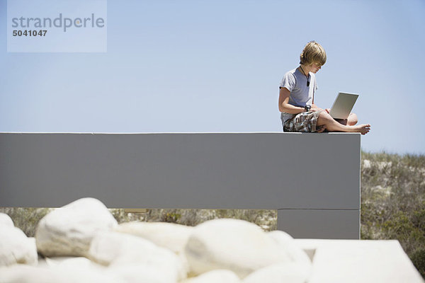 Teenager-Junge sitzt am Rande einer Terrasse und benutzt einen Laptop