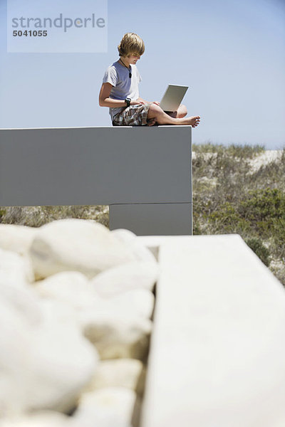 Teenager-Junge sitzt am Rande einer Terrasse und benutzt einen Laptop