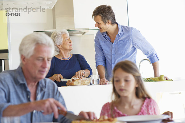 Mann beim Kuchenschneiden mit seiner Enkelin und seiner Frau im Gespräch mit ihrem Sohn im Hintergrund
