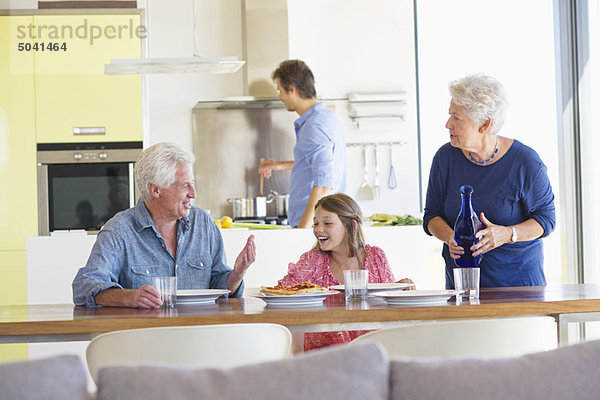 Mädchen sitzt mit ihren Eltern am Esstisch und ihr Vater kocht im Hintergrund.