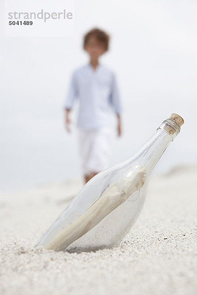 Verschwommener Junge geht auf die Flasche zu  mit einer Notiz am Strand.