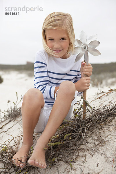 Porträt eines lächelnden Mädchens  das auf Sand sitzt und das Windrad hält.