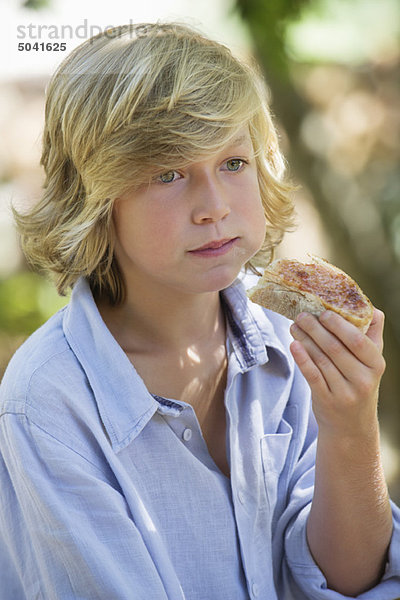 Kontemplativer kleiner Junge schaut weg  während er im Freien Brot isst.