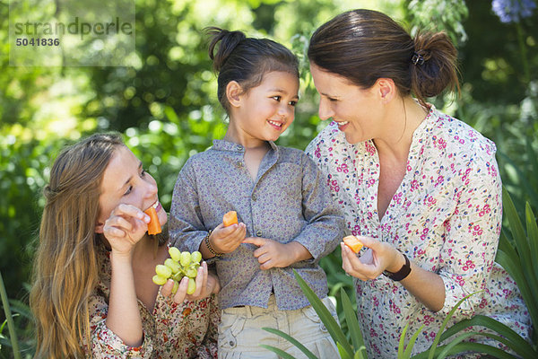 Glückliche Mutter isst Früchte mit ihren beiden Töchtern im Freien.