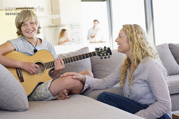 Teenager-Junge spielt Gitarre  seine Mutter sitzt neben ihm und lächelt.