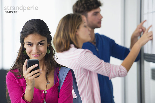 Portrait von Studenten  die ein Mobiltelefon benutzen  während Freunde im Bulletin Board nach Testergebnissen suchen.