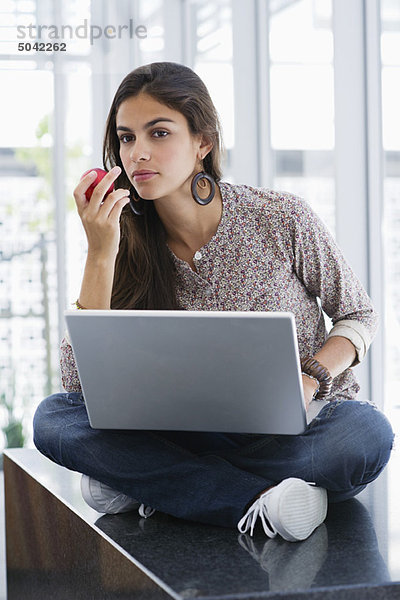 Schöne Frau  die einen Apfel isst  während sie einen Laptop benutzt.