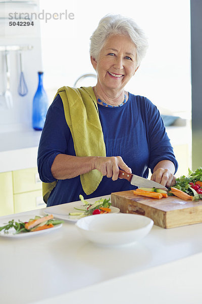 Porträt einer älteren Frau  die in einer Küche Gemüse schneidet.
