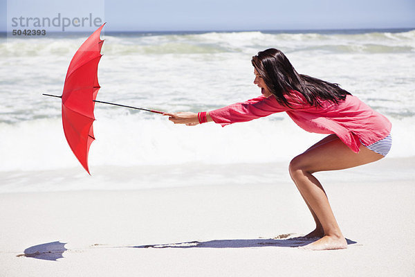 Frau  die versucht  einen Regenschirm bei starkem Wind am Strand zu halten.