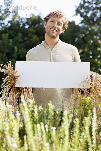 Junger Mann in Vogelscheuchenkleidern und mit einem leeren Plakat auf einem Feld.
