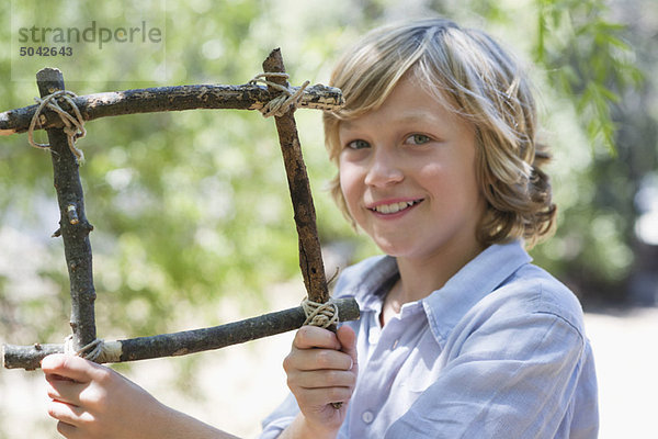 Porträt eines süßen kleinen Jungen  der einen Rahmen aus Treibholz im Freien hält.