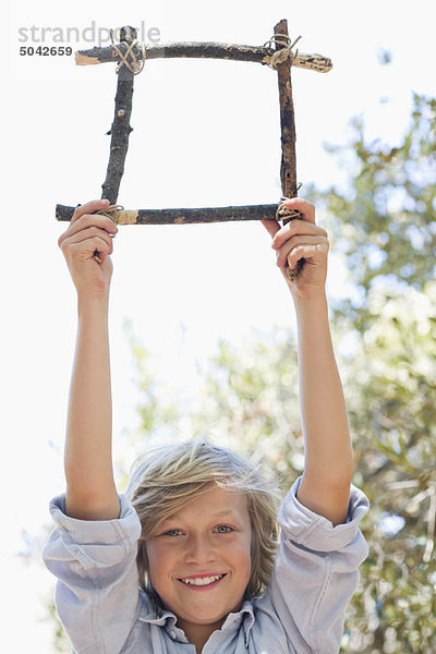 Porträt eines süßen kleinen Jungen  der einen Rahmen aus Treibholz hält  dessen Arme im Freien hochgezogen sind.