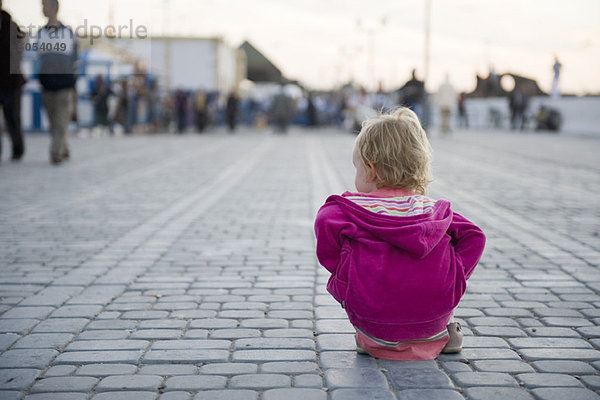 Kleinkind-Mädchen sitzt auf dem Boden auf dem Stadtplatz und sieht zu  wie Leute vorbeigehen.