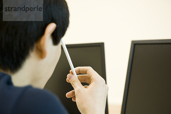 Junger Mann schaut auf Computerbildschirme  hält Bleistift gegen Kopf in Gedanken