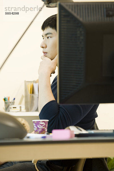 Junger Mann sitzt am Schreibtisch und schaut in Gedanken weg.