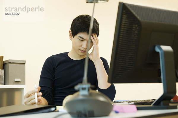 Junger Mann sitzt am Schreibtisch  hält den Kopf und schaut in Gedanken nach unten.