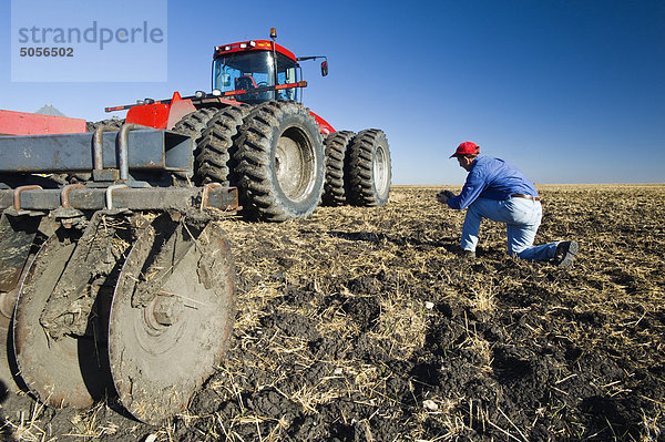 ein Mann untersucht neu kultivierten Bodens und Weizen stoppel neben einem Traktor zieht cultivating Ausrüstung in der Nähe von Lorette  Manitoba  Kanada