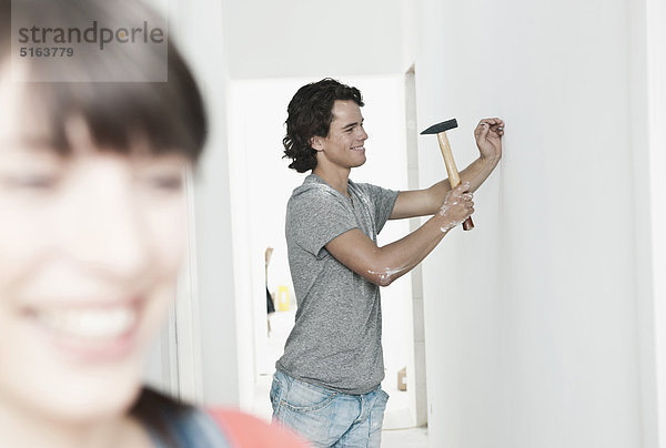 Junger Mann hämmert Nagel in renovierender Wohnung mit Frau im Vordergrund