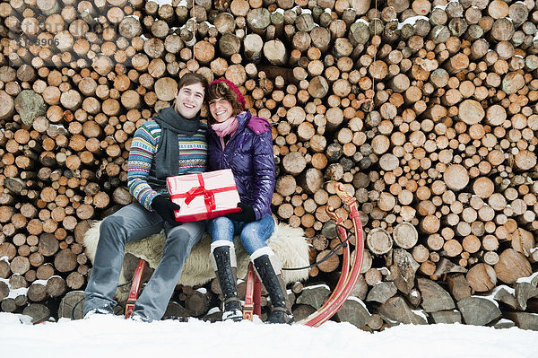 Österreich  Salzburger Land  Flachau  Junger Mann und Frau auf Schlitten sitzend mit Weihnachtsgeschenk und Brennholz im Hintergrund
