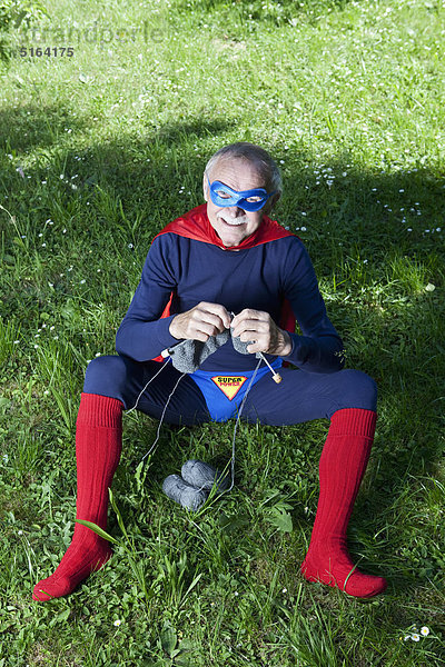 Österreich  Burgenland  Senior in Superman-Kostüm auf Gras sitzend und strickend