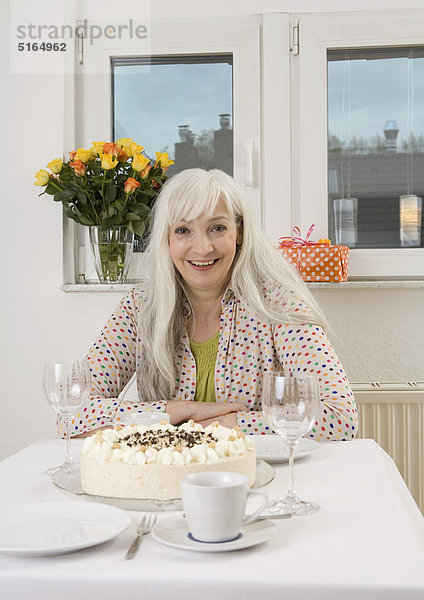 Frau am Esstisch sitzend mit Kuchen  Flowervase und Geschenk  lächelnd  Portrait