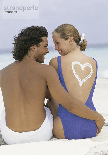 Paar  sitzen am Strand  sie hat Herz aus Sonnencreme auf Rücken