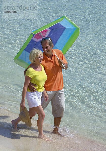 Älteres Paar geht mit Sonnenschirm am Strand spazieren