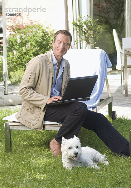 Mann sitzt mit Laptop auf Gartenliege  Hund liegt im Gras