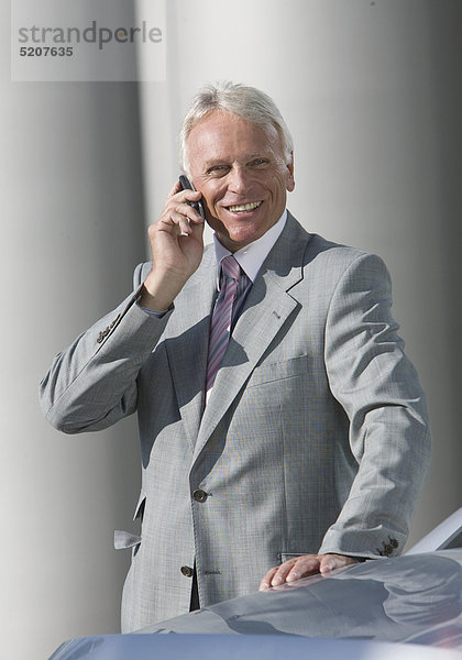 Älterer Mann steht im Anzug neben Auto  telefoniert mit Handy
