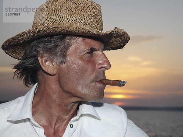 Älterer Mann mit Strohhut und Zigarre vor Sonnenuntergang am Meer