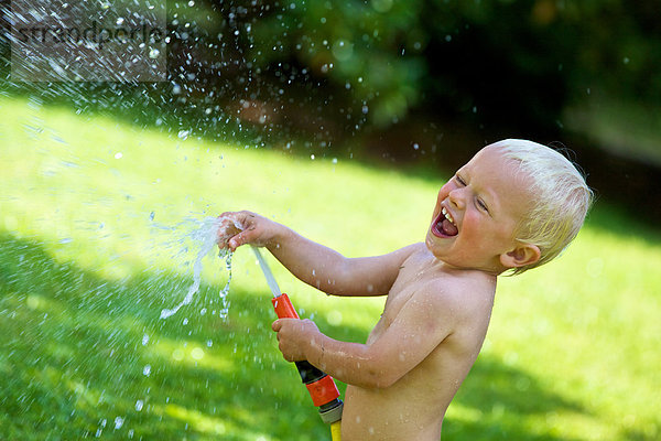 Kleiner blonder Junge spritzt mit Wasser im Garten