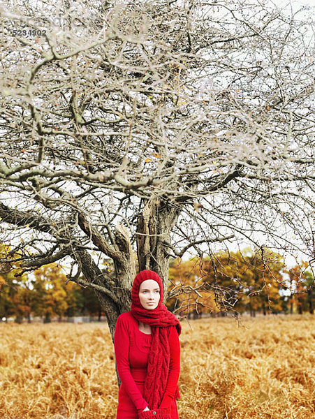 Eine Frau in einem leuchtend roten Kleid und Schal unter einem blattlosen Baum in der Natur.