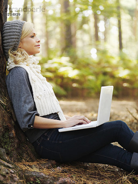 Eine Frau in der Natur  die sich an einen Baum lehnt und einen Laptop benutzt.