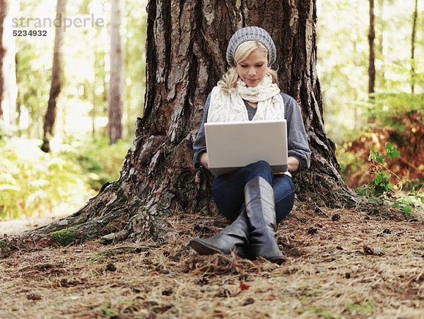 Eine Frau in der Natur  die sich an einen Baum lehnt und einen Laptop benutzt.