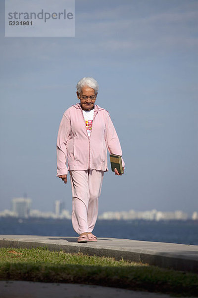 Eine ältere Frau  die mit einem Buch auf einem Weg am Meer spazieren geht.