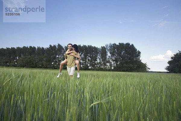 Ein Mann  der seine Freundin huckepack durch ein Weizenfeld fährt.