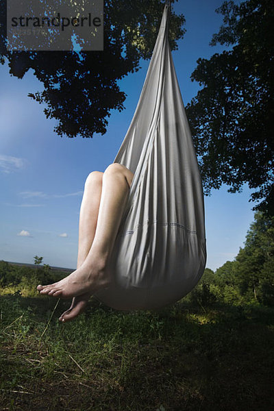 Eine Frau sitzt in einer geschlossenen Hängematte  die an einem Baum hängt  nur die Beine sind sichtbar.