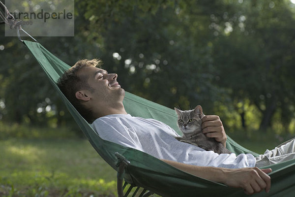 Ein Mann  der in einer Hängematte liegt und eine Katze streichelt.