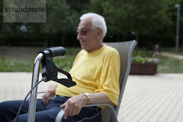 Ein älterer Mann auf einem Stuhl sitzend  der Griff seines Gehers im Vordergrund  konzentriert sich auf den Vordergrund.