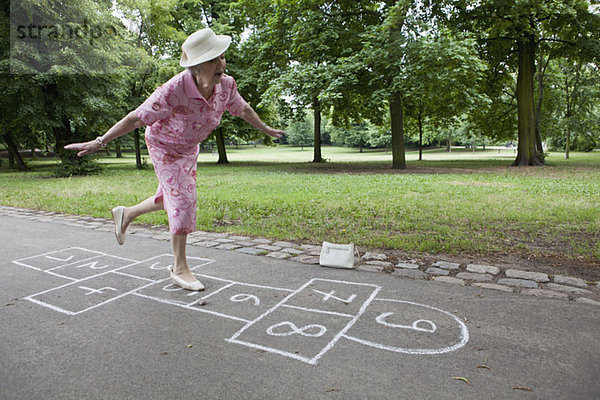 Seniorin spielt Hopscotch und versucht  ihr Gleichgewicht zu halten.