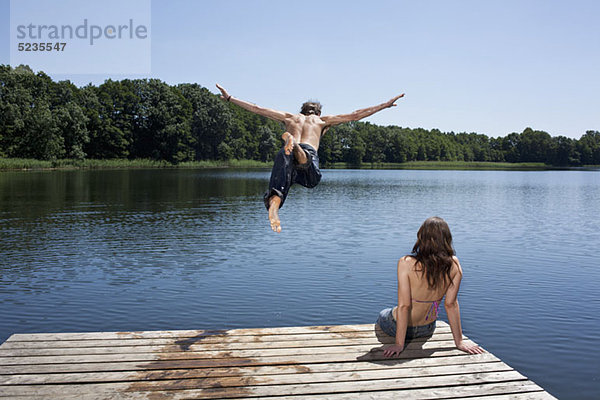 Der Typ taucht mit ausgestreckten Armen in den See  während das Mädchen am Steg zuschaut.