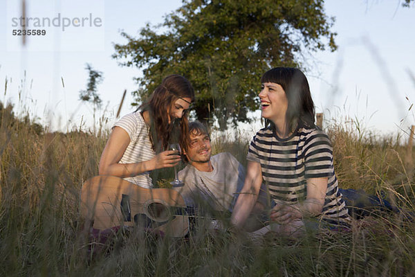 Drei Freunde genießen das Sitzen auf einem abgelegenen Feld mit Wein und Gitarre.
