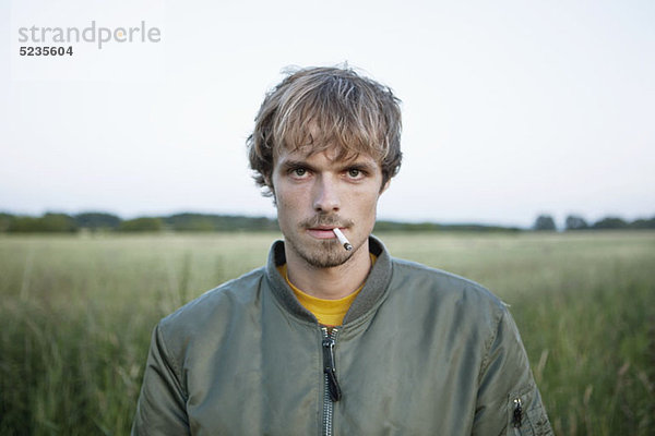Profil eines Mannes  der auf einem abgelegenen Feld steht und eine Zigarette aus dem Mund hängt.