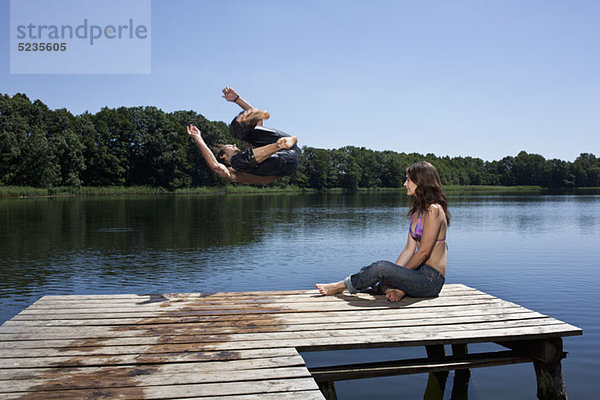 Der Typ springt mit ausgestreckten Armen in den See  während das Mädchen am Steg zuschaut.