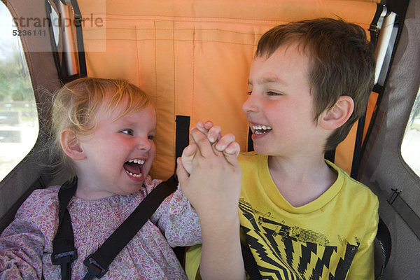 Bruder und Schwester reiten zusammen im Kinderwagen  lachend und Händchen haltend.
