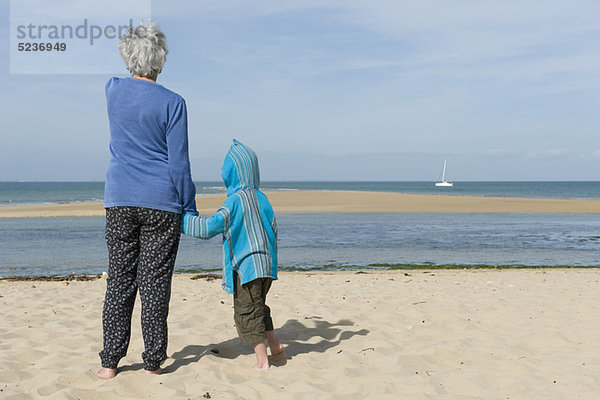 Großmutter und Enkel stehen zusammen am Strand und sehen das Boot am Horizont.