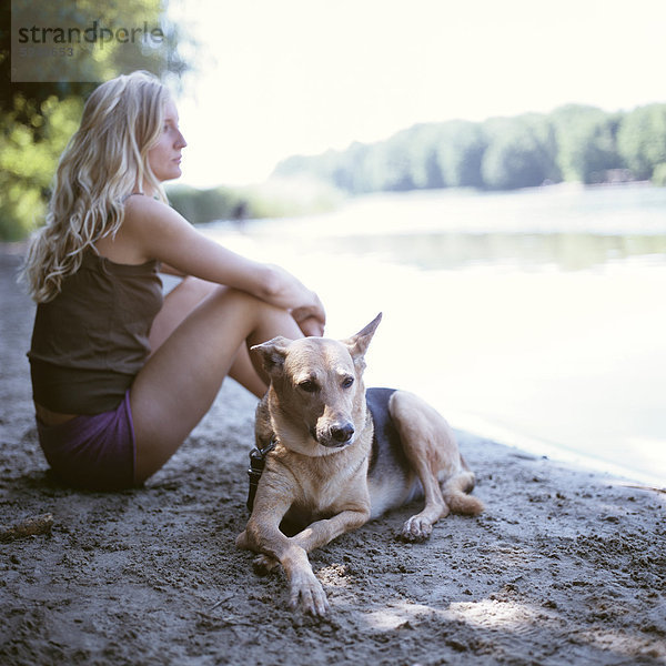 Frau sitzt mit Hund an Seeufer