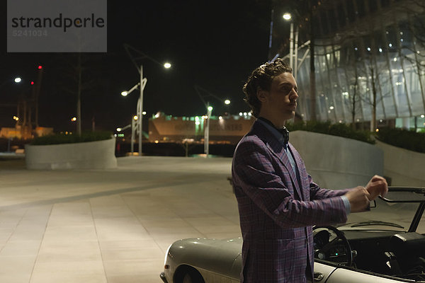 Mann  der nachts neben einem klassischen Cabriolet steht.