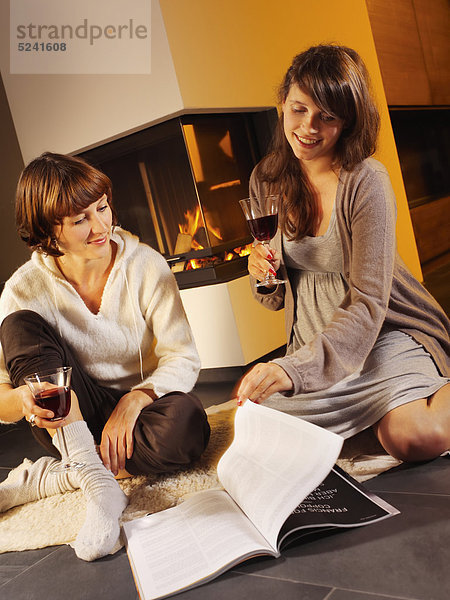 Zwei junge Frauen schauen vor Kaminofen Zeitschrift an  trinken Rotwein