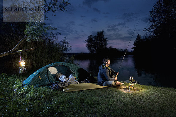 Mann sitzt am Seeufer mit Laterne und fischt in der Abenddämmerung.